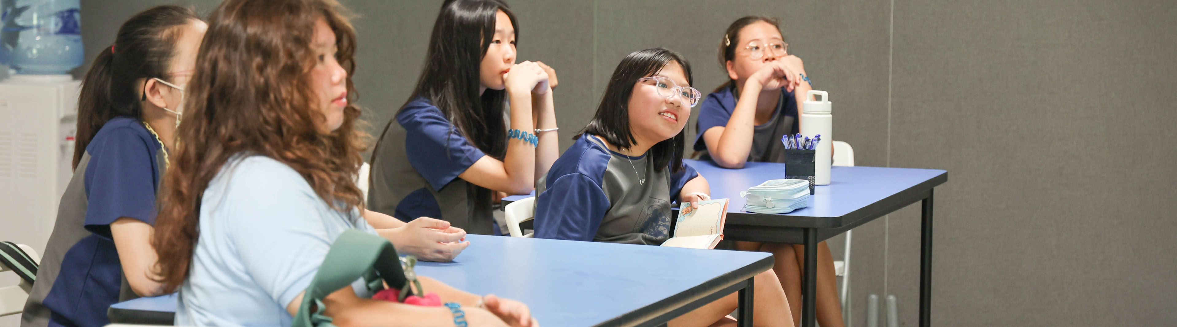 A group of students sitting at desks in a classroom, looking towards the front.